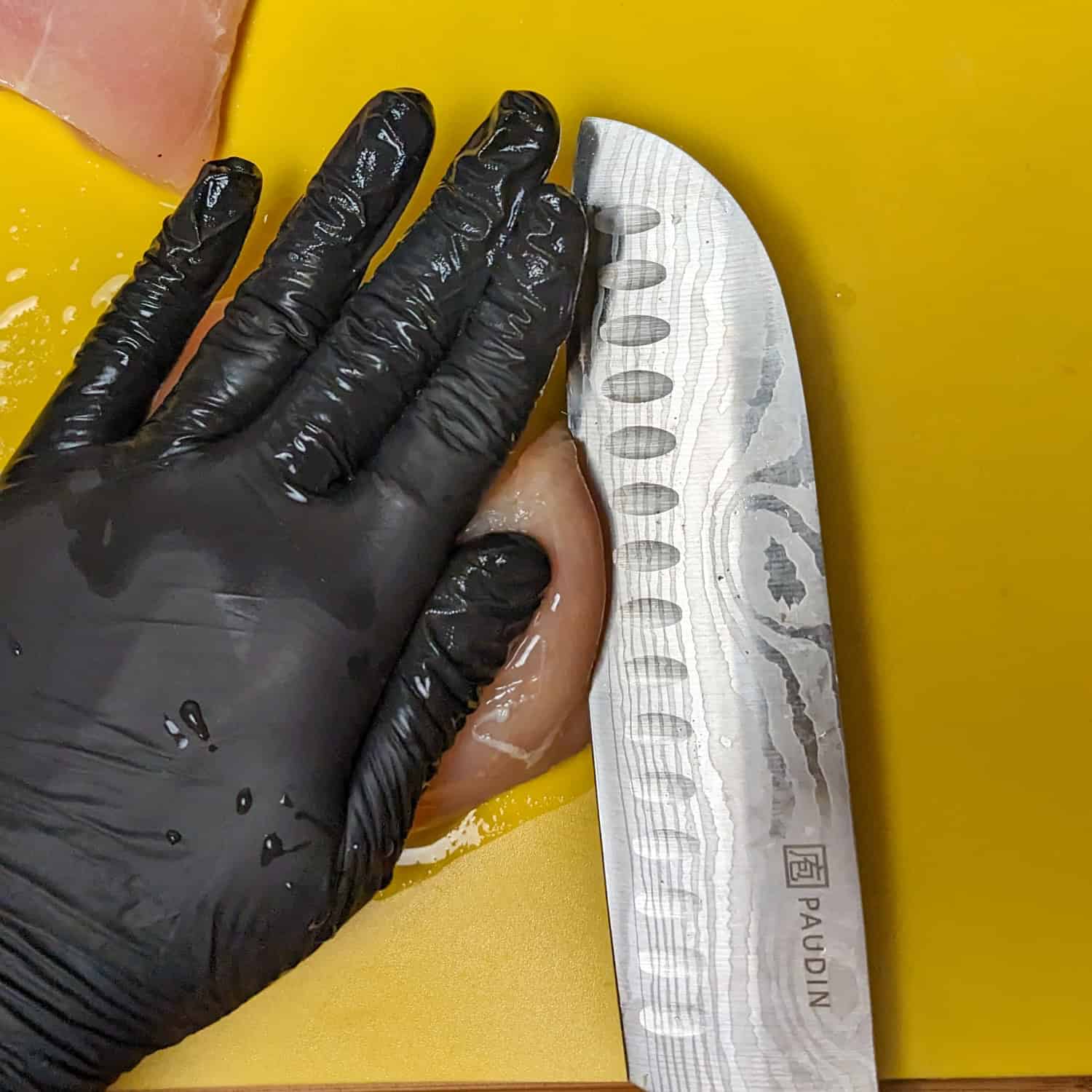 gloved hand over a chicken breast while a knife slices the it in half on a plastic yellow cutting board.